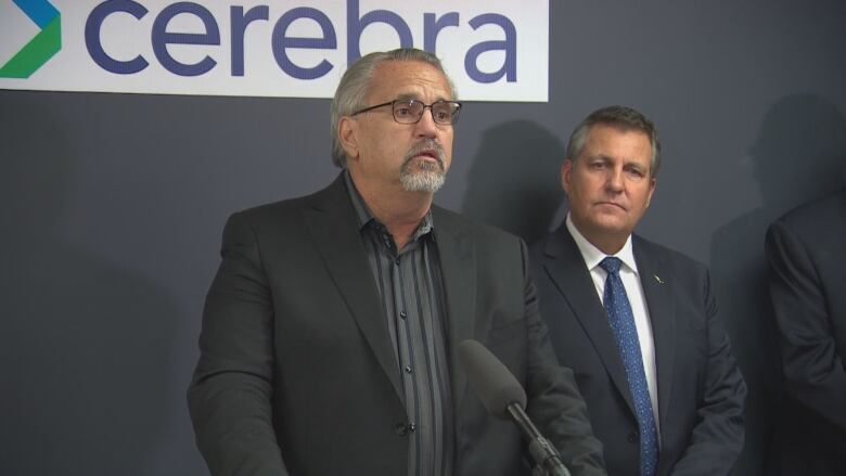 A man in a suit with glasses and a short grey beard speaks into a microphone as another man in a suit stands behind him.