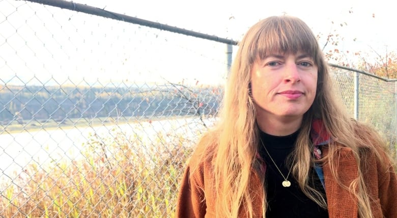 A woman in a brown jacket stands by a chainlink fence with a river running behind it.