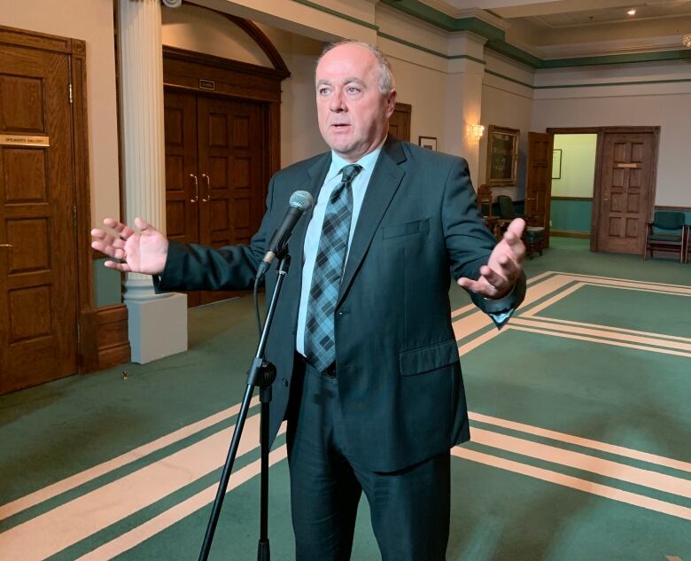 A person wearing a suit stands while speaking into a microphone. The person is gesturing with their arms outstretched.