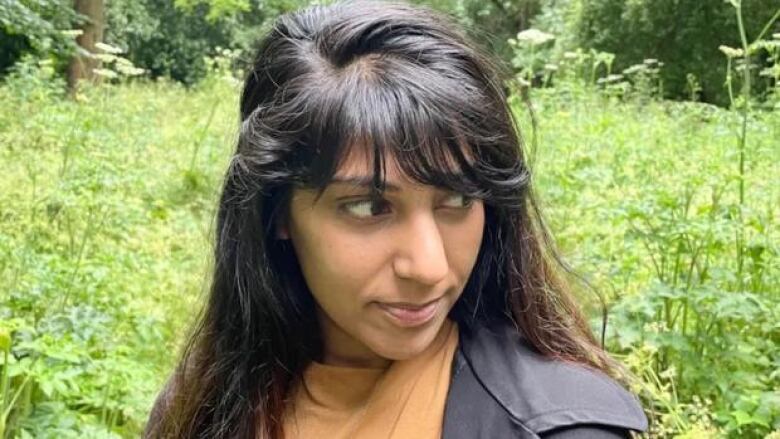 Portrait of the writer with long black hair and wearing an orange shirt and black jacket while standing in a field of leafy green plants