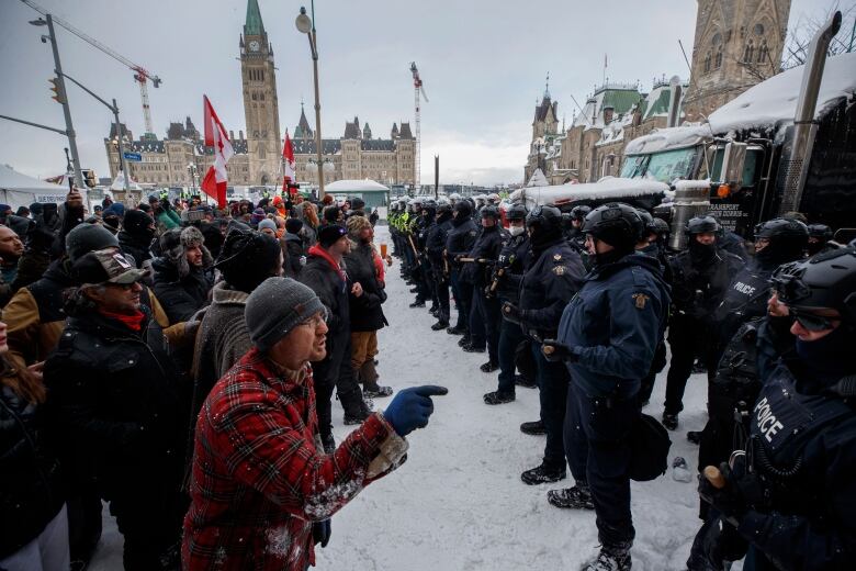 A row of protesters yells at a row of police officers in front of a legislature in winter.