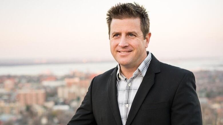 A person in in a plaid shirt and blazer smiles in front of a view from on top of the Niagara Escarpment