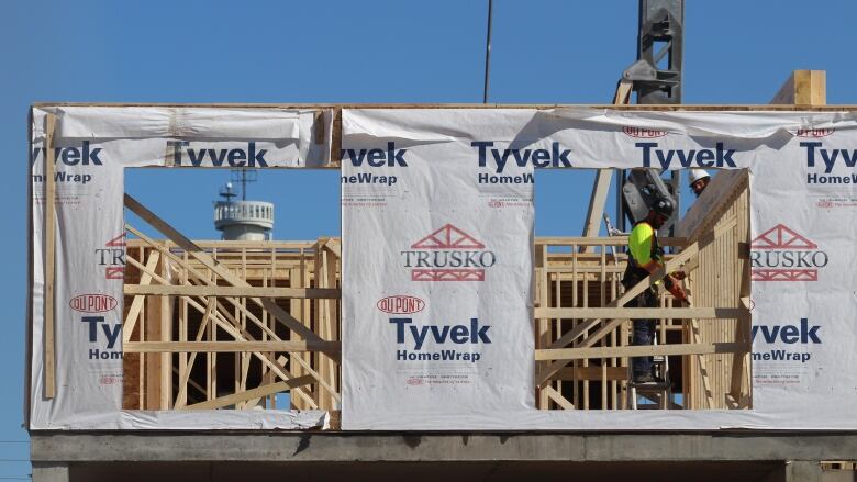 A worker wearing a hard hat stands inside a building in the framng phase of construction. 