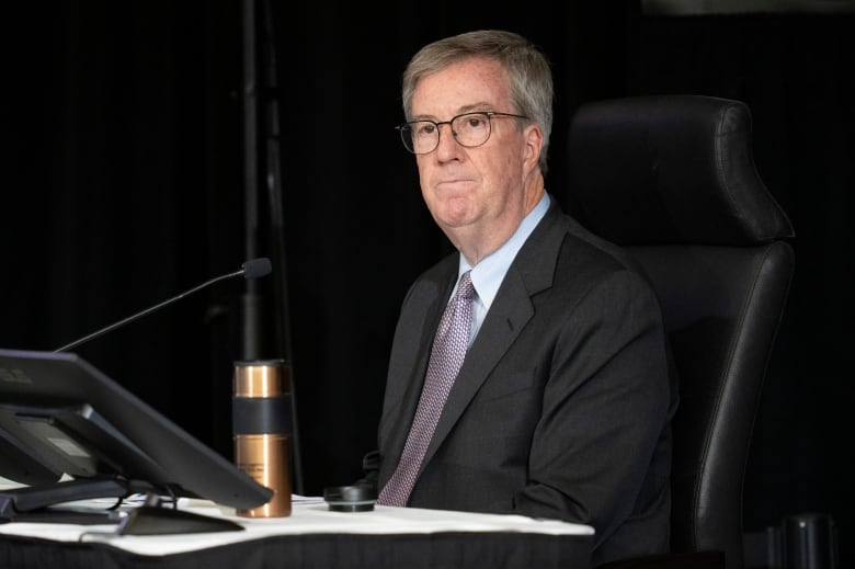 A politician looks at a room from behind a desk with a microphone and water bottle on it.