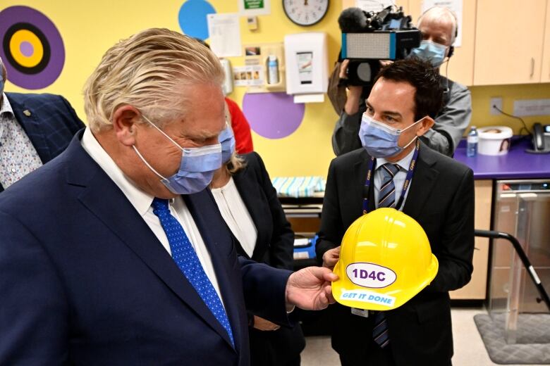 One man in a suit passes a yellow hard hat to another.