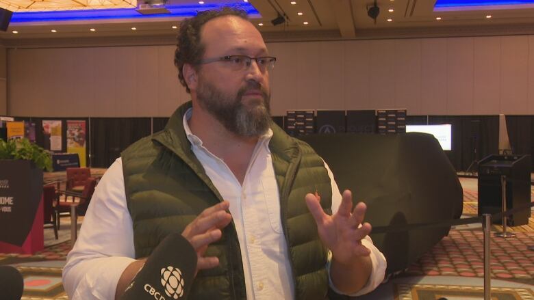 Flavio Volpe speaking to reporter, wearing green vest and glasses. 