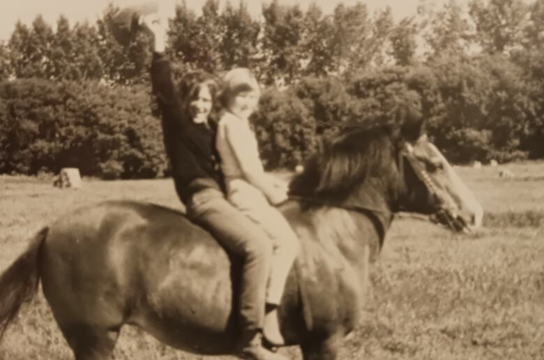 A black and white photo featuring two children sitting atop a dark horse. One child waves at the photo-taker. 