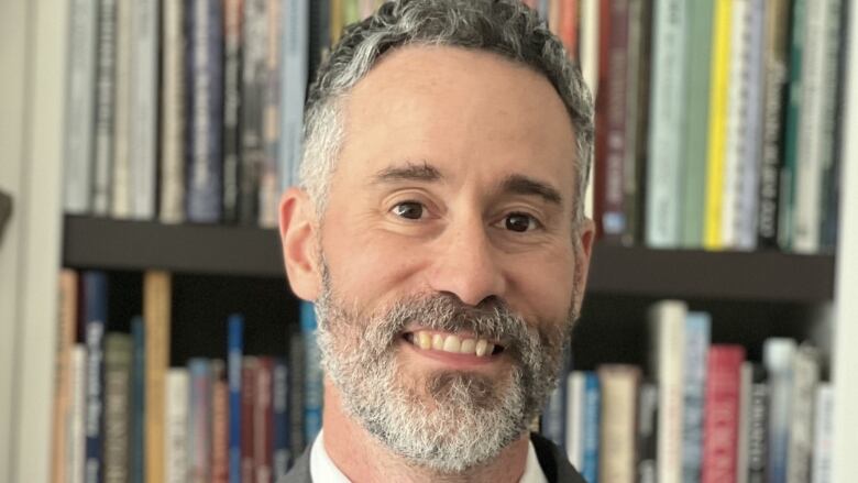 A close up headshot of a person with a beard in front of books.