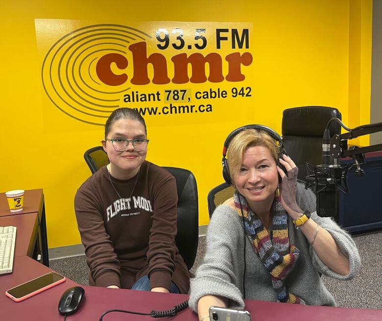 A mother and daughter sit behind the microphone in a radio studio. The walls are a bright yellow, and the back wall features the logo for CHMR 93.5 FM.