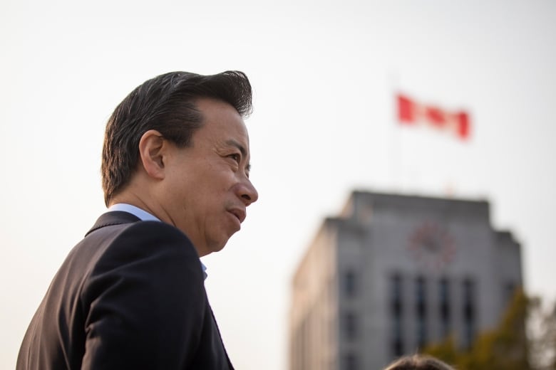 A man stands in profile with Vancouver city hall and a Canadian flag in soft focus to the right.