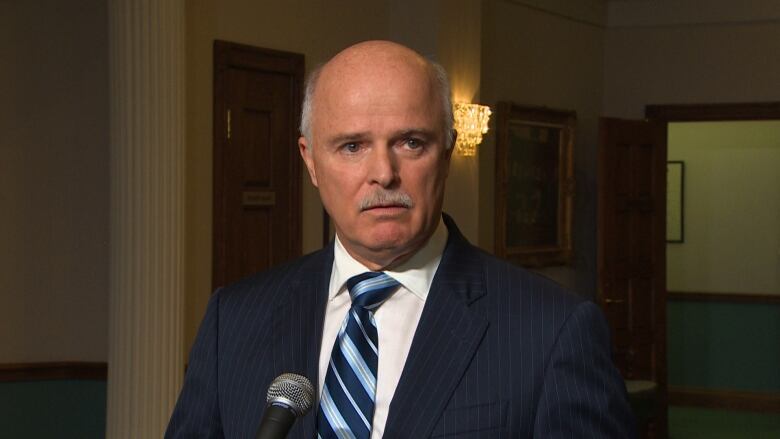 A man wearing a navy suit with a blue tie speaks with reporters in Confederation Building.