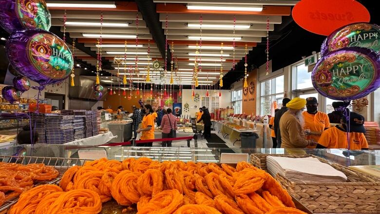 A store is decorated for Diwali, with gold tassles hanging from the ceiling and purple helium balloons floating that say 'Happy Diwali'. More than a dozen shoppers navigate through stacks of sweets and boxes. In the front of the photo is a tray of twisted deep fried dough dipped in sugar syrup that's a bright orange colour. 