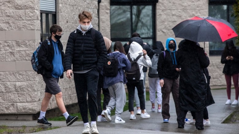 Students wearing masks walk in the rain outside a school building. 