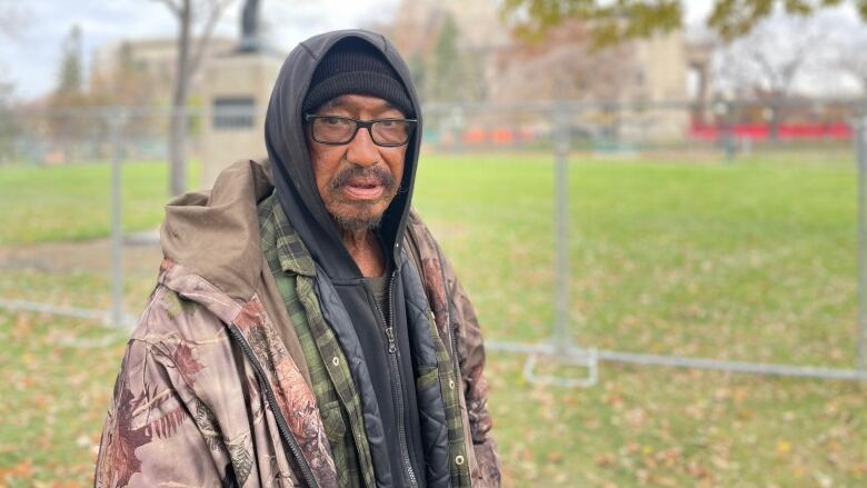 A man wearing a camouflage jacket, a hoodie and glasses stands in a grassy area with a large domed building in the background behind him.