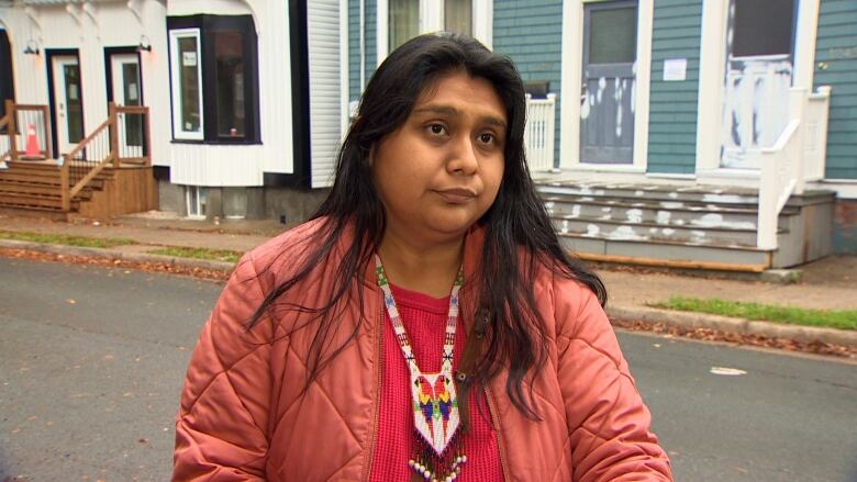 A woman with long black hair and a pink jacket stands on a residential street with houses in the background.