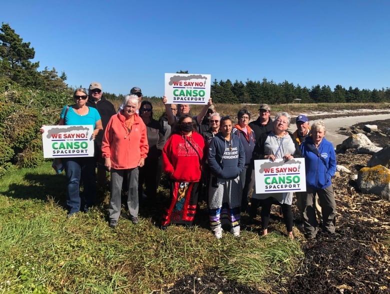 16 people, three holding signs, stand on a beach