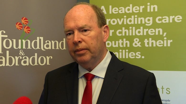 A middle-aged man wearing a black suit with a red tie speaks with reporters. He stands in front of a banner showing Newfoundland and Labrador's logo, along with a logo from Waypoints that reads 'A leader in providing care for children, youth & their families.'