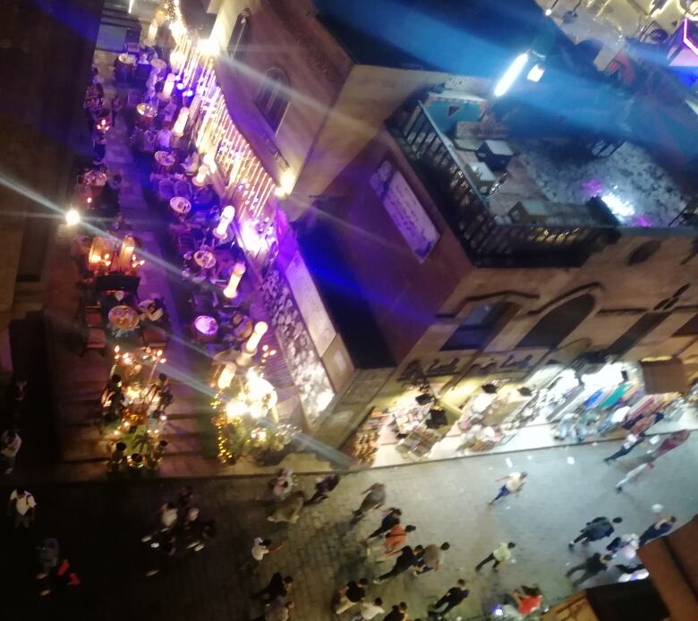 A street with people walking and dining outdoors is surrounded by lights. 