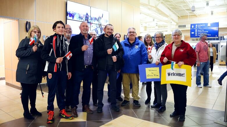 A group of people smile at the camera while holding Canadian and Ukrainian flags and homemade signs saying 'Welcome.' 