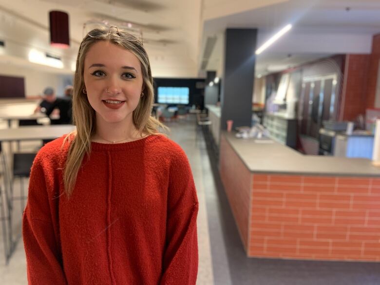 A waste-up shot of a person with long blonde hair wearing a red sweater. To their left, out of focus, is a grey bar.