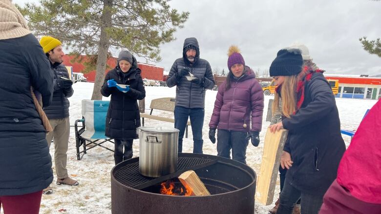 A group of people stand around a fire pit with a large pot of soup.