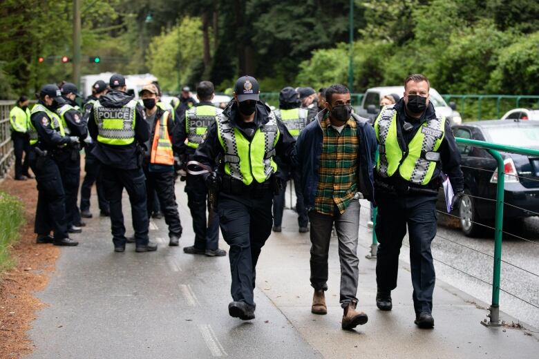 A man wearing a mask walks with his hands behind his back, between two police officers. 