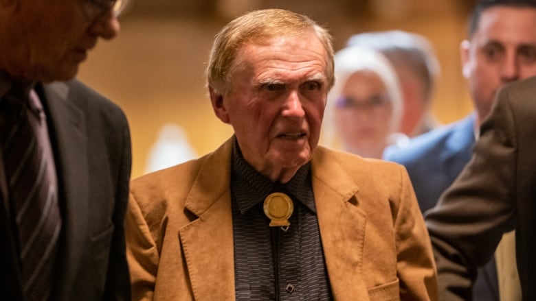 A man in a brown jacket and bolo tie stands among other people.