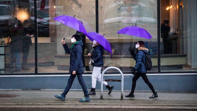 Three people walk past a storefront with purple umbrellas.