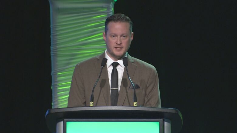 A man in a dark suit stands at a green-lit podium.