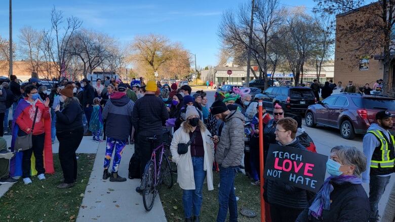 Dozens of people are seen standing in a grassy area beside a city street, including one holding a sign reading 