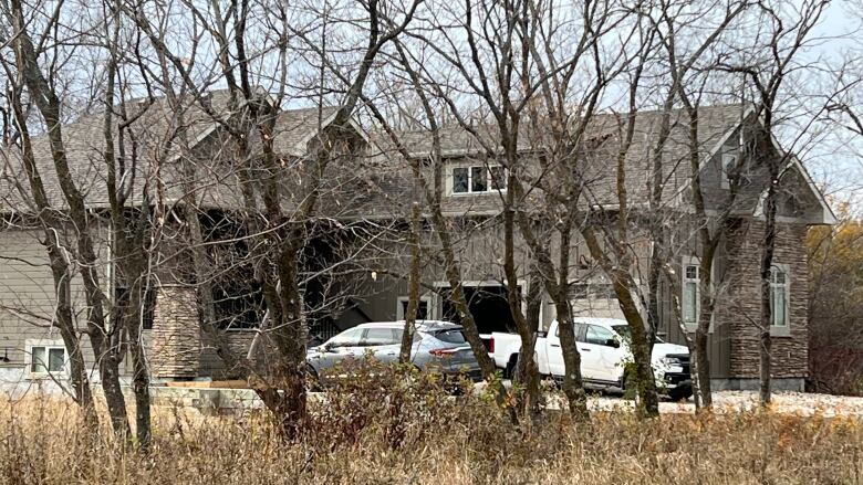 A house with two vehicles parked in the driveway is seen through trees whose leaves have fallen.