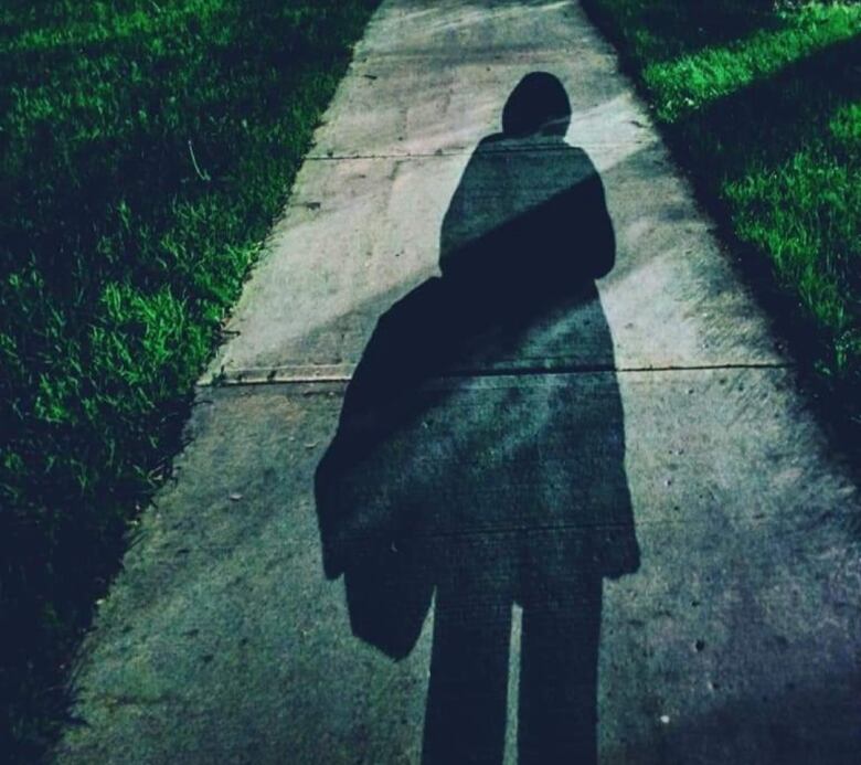 Shadow of the back of a woman, wearing a heavy coat and carrying a tote bag, walking down a sidewalk.