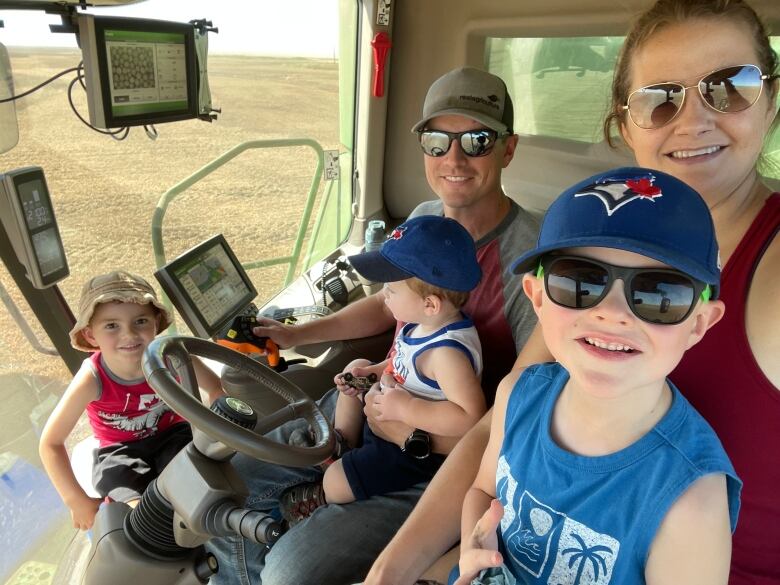 A man operates a combine on a golden field. He has a small child on his lap. A woman sits next to him with a child on hers, and another young boy sits on the floor of the cab by the wheel.