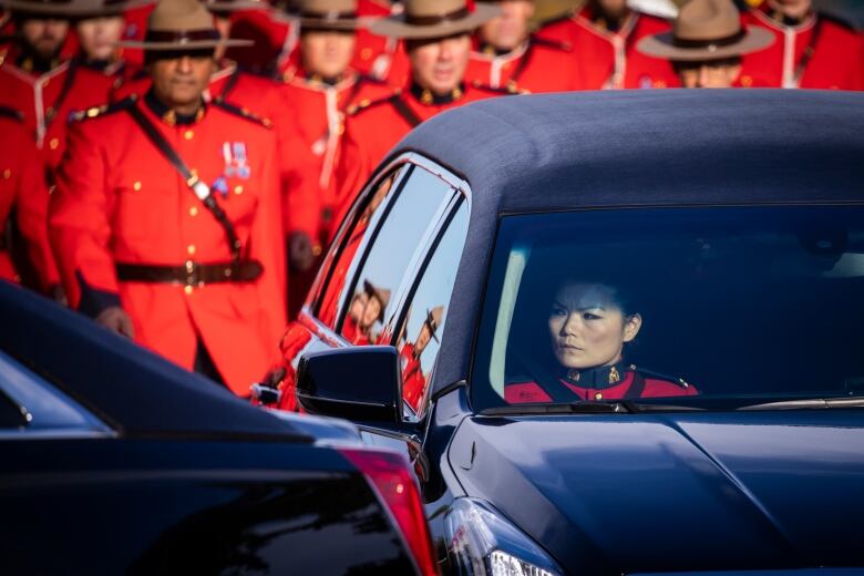 A young Asian female Mountie in red serge sits in a black vehicle staring out the window with a parade of RCMP officers behind her also dressed in red serge.