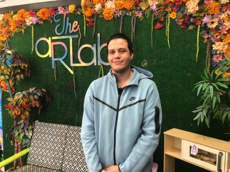 A man wearing a light blue zipper hoodie standing in front of a colourful sign that says 'The Art Lab'