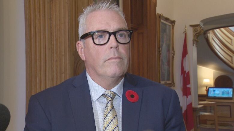 Man in centre of screen wearing glasses, blue suit, patterned tie and a red poppy on his suit jacket. There is a collapsed Canadian flag behind him. 