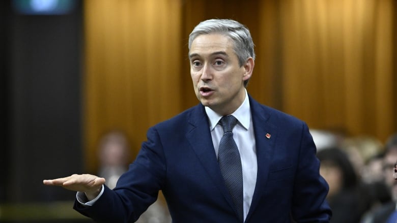 A man with white hair and a blue suit gestures with is right hand.