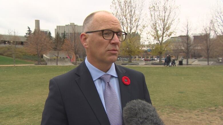 A man in a suit with a poppy on his lapel speaks into a micrphone in the middle of a field.