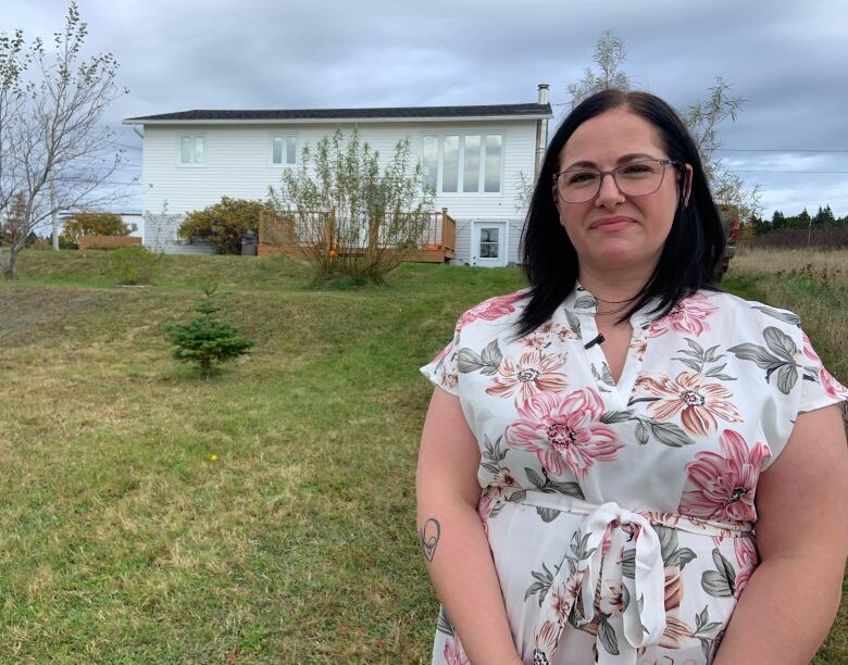 A waist-up shot of a person wearing a floral blouse stands outside in the foreground. A house can be seen in the background to her left, along with some small trees.