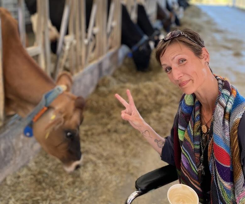 A woman holds up fingers in peace sign beside cow eating hay.