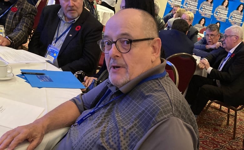 A man wearing glasses and a short-sleeved shirt sits at a round table during a conference.