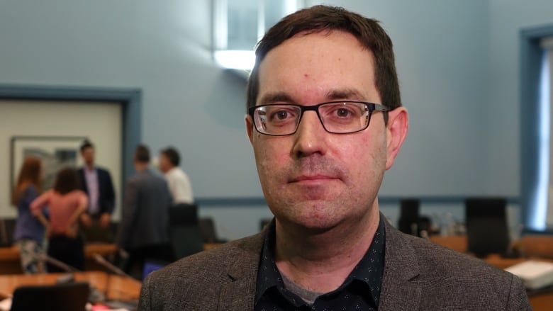 A man in black glasses and a blazer with dark hair stands in front of a round table