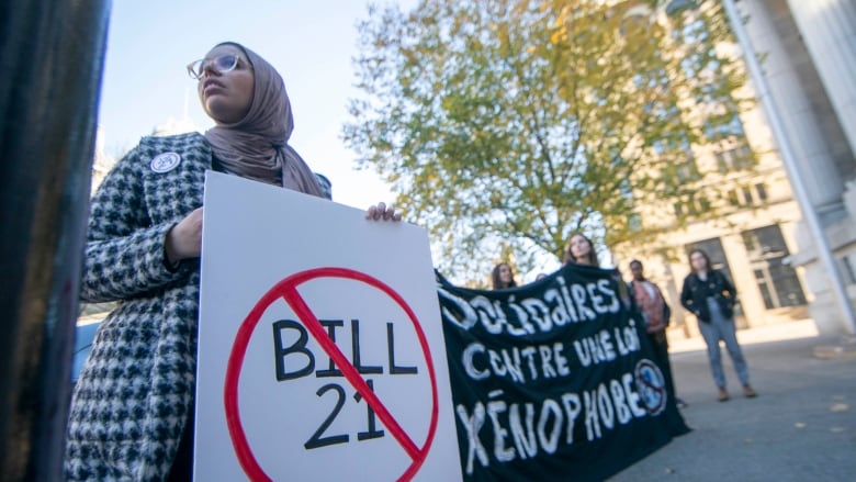 A woman in a hijab holds a sign with Bill 21 crossed out. Others hold a black banner.