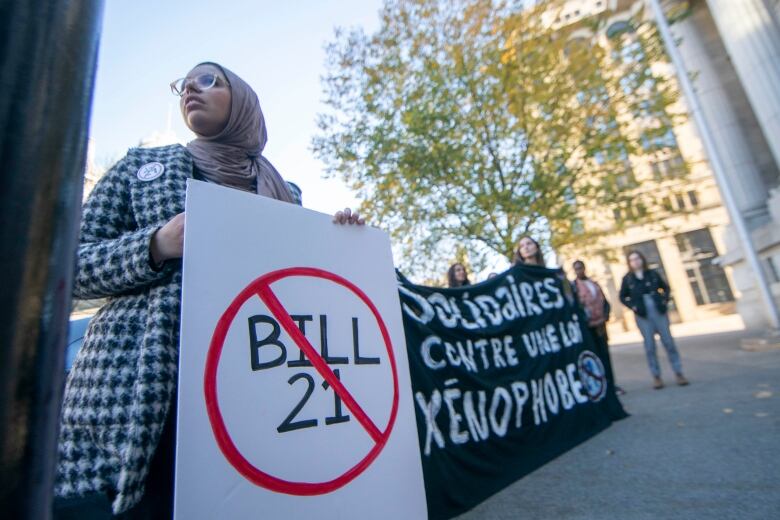 A woman in a hijab holds a sign with Bill 21 crossed out. Others hold a black banner.