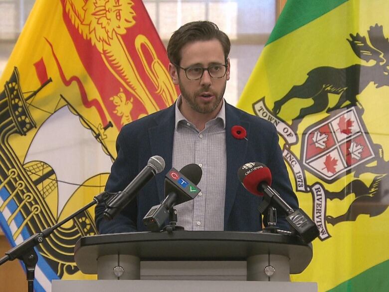 A man soeaking at a podium with the New Brunswick flag and the Town of Riverview flag behind him.