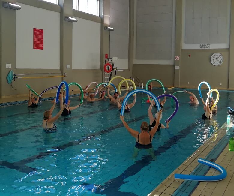 A group of people are in a shallow pool lifting pool noodles over their heads as part of a water fitness class. 