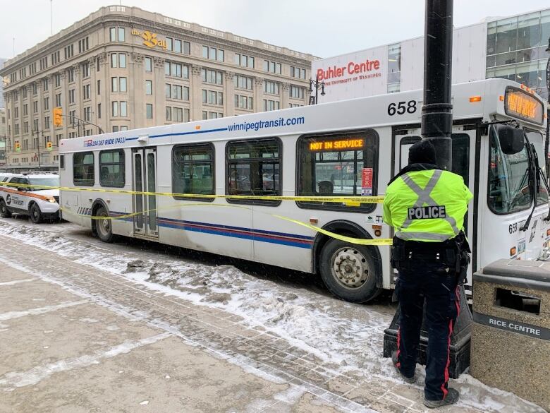 White bus on the street in front of the Buhler Centre on Portage Avenue, with a police officer securing yellow police tape to a lamp post beside it.