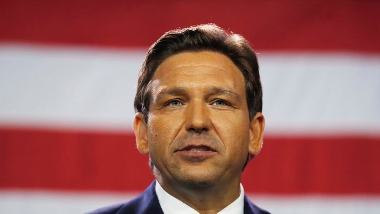 A man with short brown hair, wearing a navy blue suit with a red tie, visible from the shoulders up, in front of the red and white stripes of a large American flag.