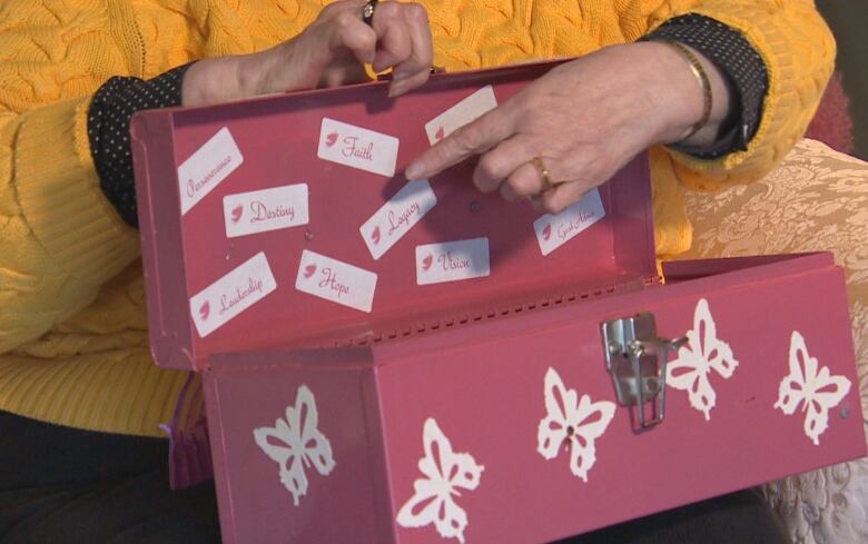 Close up of an open pink metal toolbox with white butterfly stickers on the outside. A hand points to individual stickers inside the toolbox that say: Perseverance. Leadership. Faith. Destiny. Hope. Voice. Legacy.