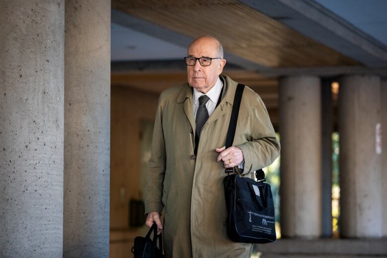 An older man with black-framed glasses in a khaki trench coat carrying a bag on his shoulder and one in his hand walks outside a Surrey, B.C. courtroom.